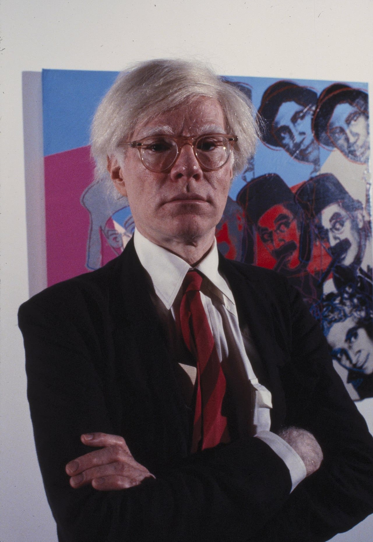 Andy Warhol standing with arms crossed, in front of his artwork, wearing glasses and a suit, photo by Bernard Gotfryd at the Jewish Museum.