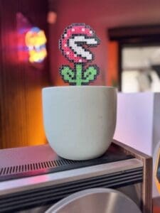 A pixel art Piranha Plant from Super Mario Bros. in a white ceramic pot, photographed in RIOT Studios with colorful neon lights in the background.