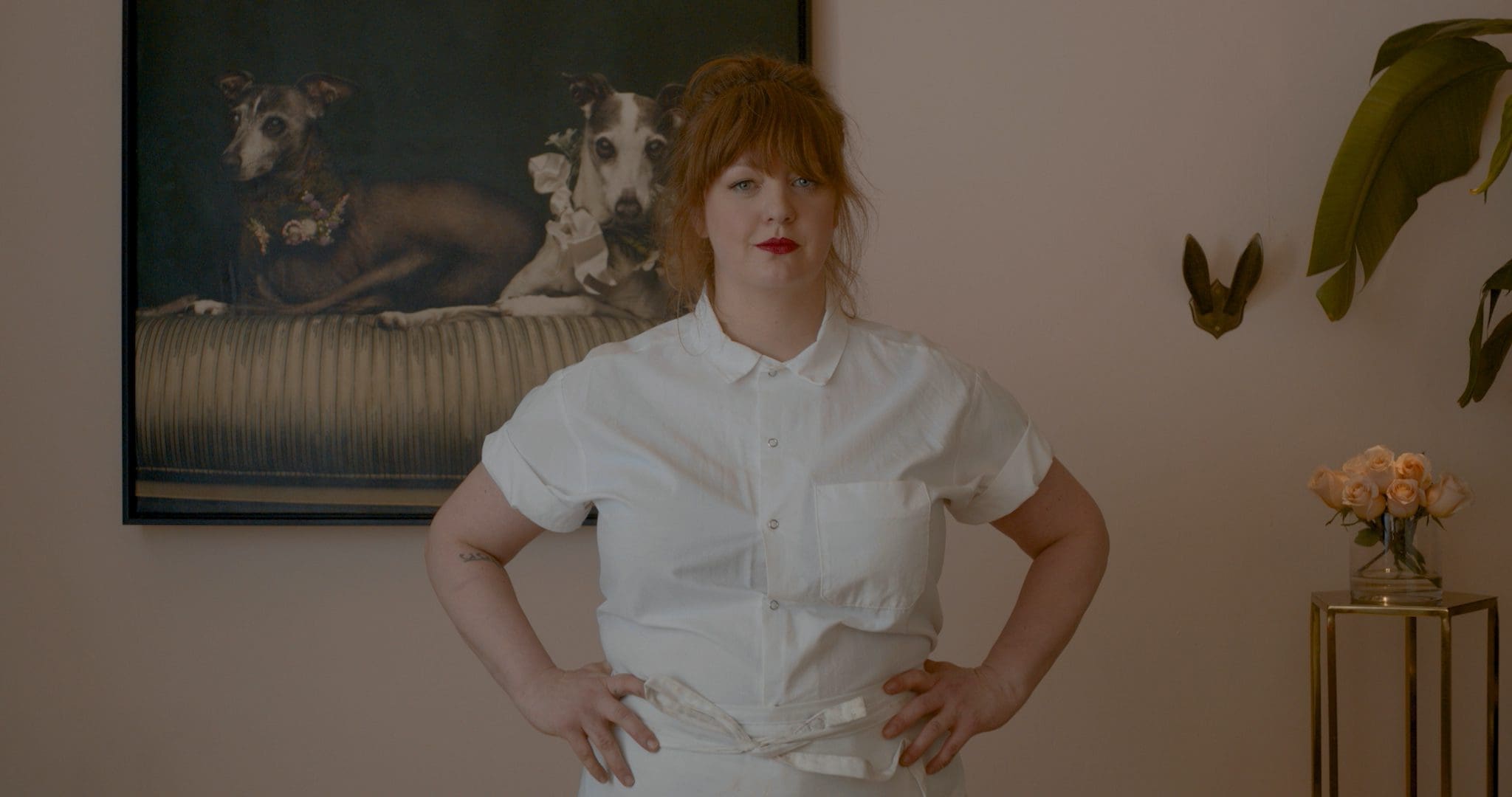RIOT, NYC Creative Agency: Chef Jamie Malone standing in front of a framed portrait of her beloved dogs, showcasing her connection to home and her personal life. Chef Jamie Malone standing in front of a framed portrait of her beloved dogs, showcasing her connection to home and her personal life.