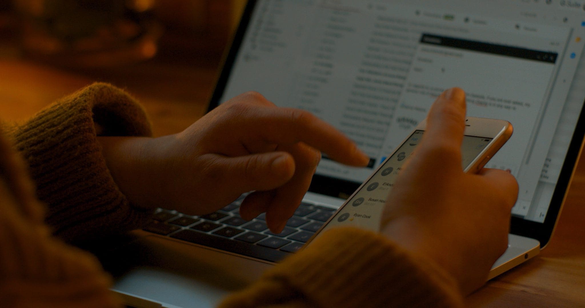 RIOT, NYC Creative Agency: Close-up of Jamie Malone working in a cozy home office, using a laptop and smartphone, showcasing multitasking and a warm, productive space. Close-up of Jamie Malone working in a cozy home office, using a laptop and smartphone, showcasing multitasking and a warm, productive space.