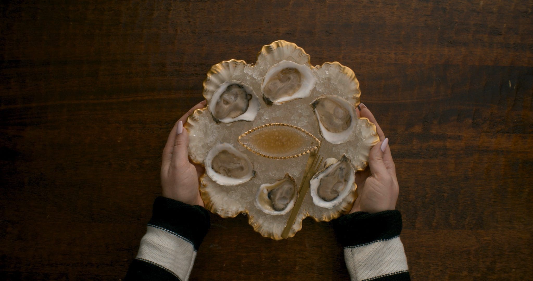 RIOT, NYC Creative Agency: A beautifully presented platter of oysters on ice, held by hands, with golden accents and a vintage feel, showcasing Jamie Malone's culinary artistry. A beautifully presented platter of oysters on ice, held by hands, with golden accents and a vintage feel, showcasing Jamie Malone's culinary artistry.