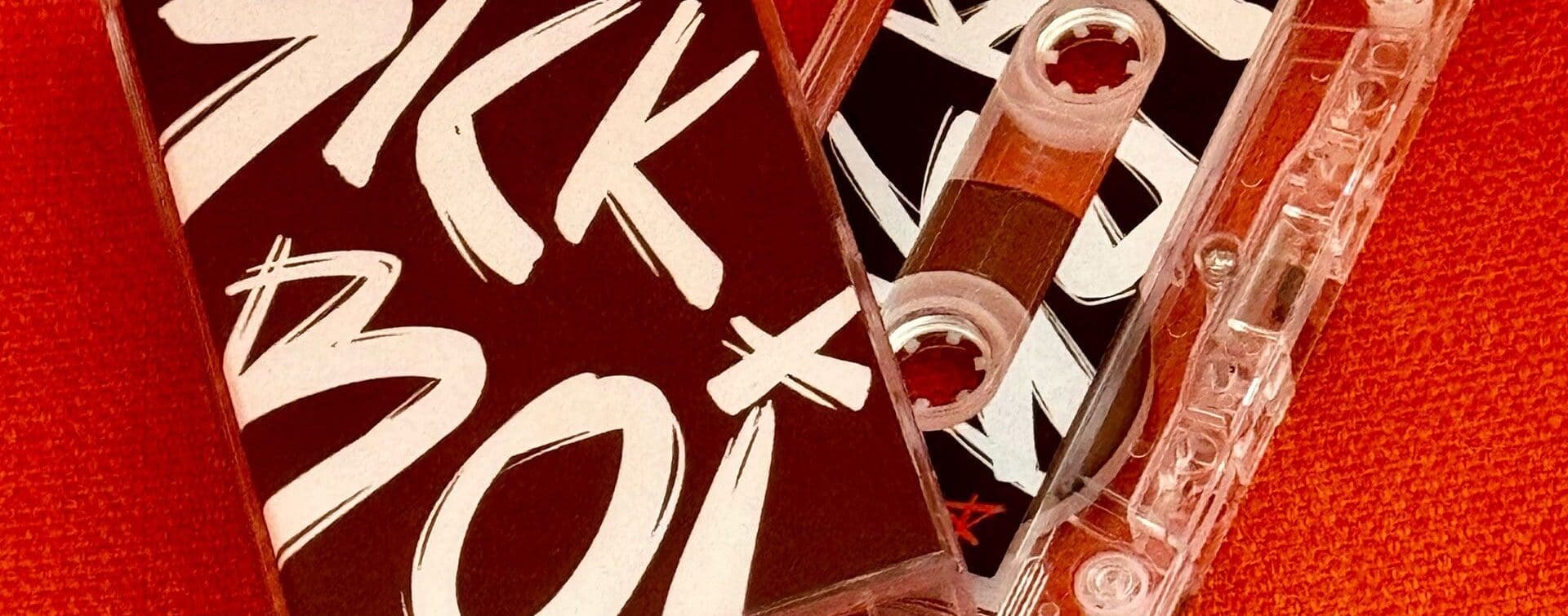 Cassette tape of Ren's 'Sick Boi' album with bold lettering on the cover, placed against a vibrant orange background.