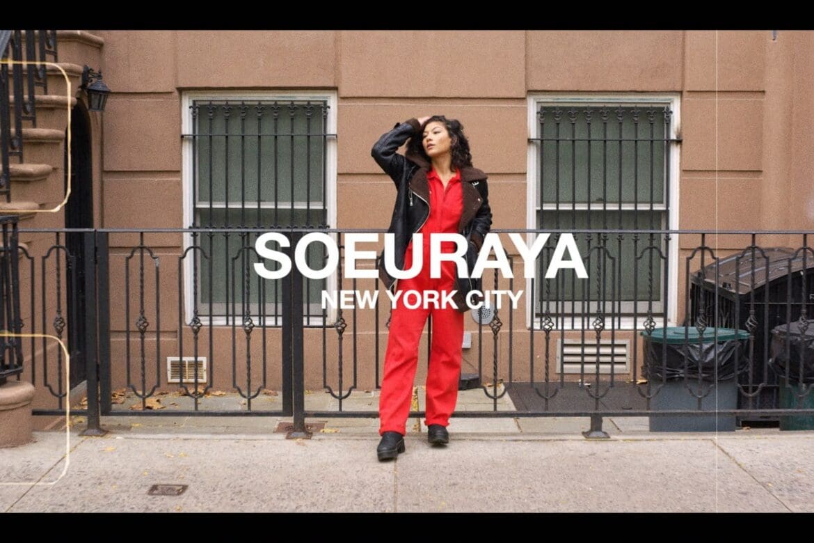 Soureya, poised and stylish in red jumpsuit and black jacket, stands confidently on a New York City street, embodying the urban energy and personal empowerment central to the SoulCycle Docufilm narrative