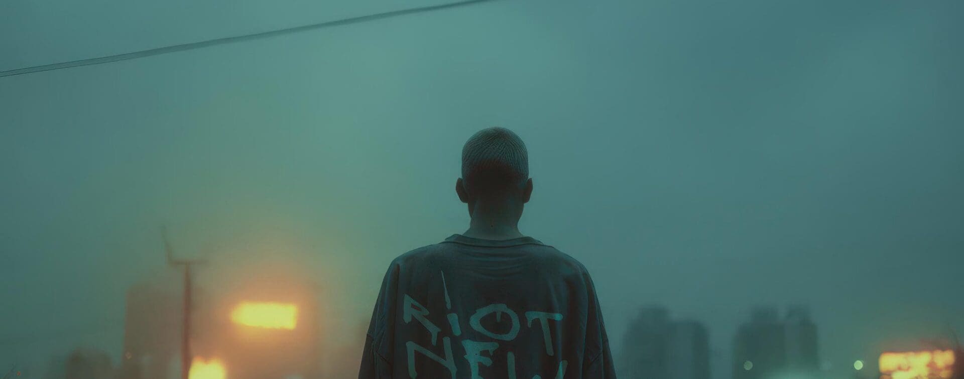 Person in oversized “RIOT NEW YORK” tee facing a rainy neon city at dusk