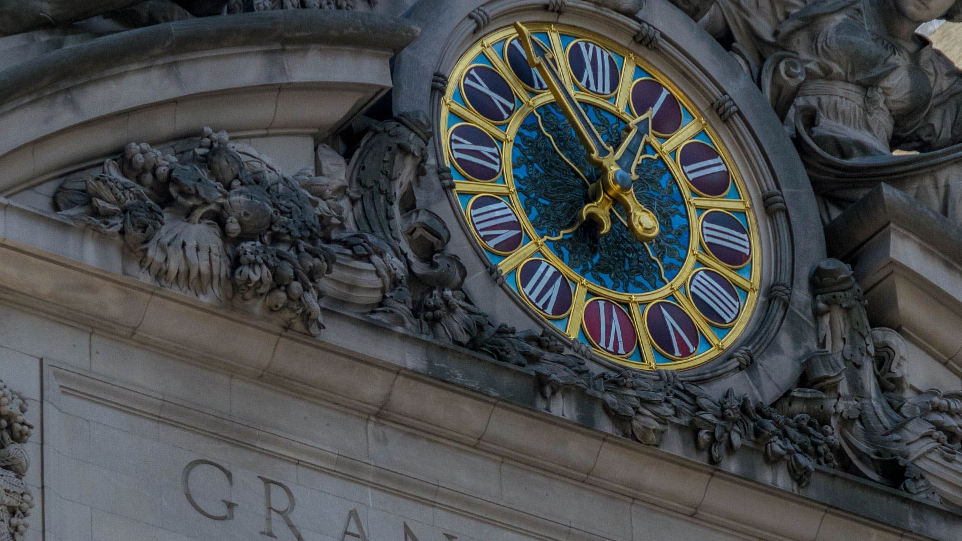 Close-up of Grand Central Terminal’s Tiffany clock face, showing ornate Roman numerals and decorative sculptures. Cropped image from Wikimedia Commons.