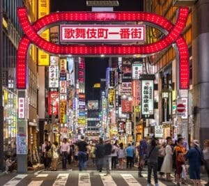 The illuminated red Kabukichō gate and neon-lit street signs at night in Shinjuku, Tokyo, with crowds of people walking beneath it.