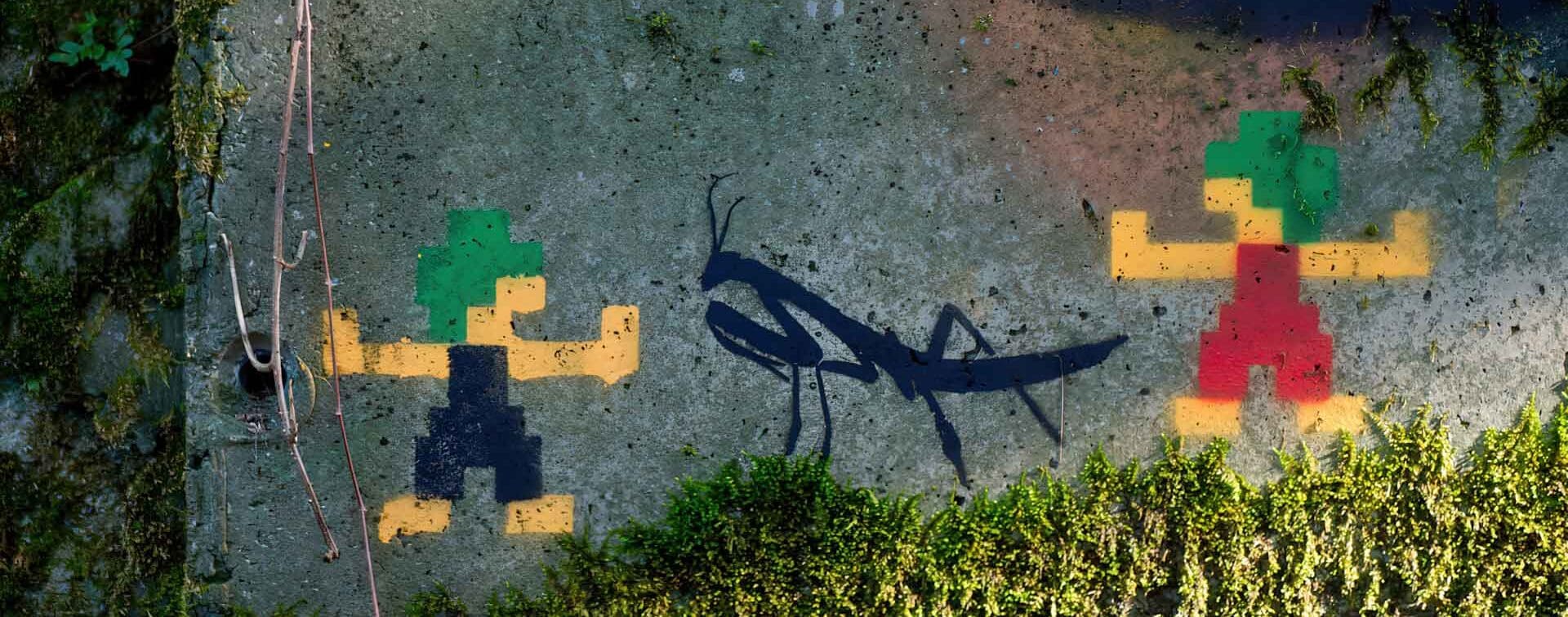 Pixel-style graffiti of Lemmings characters on a moss-covered concrete wall, with a black silhouette of a praying mantis between them.