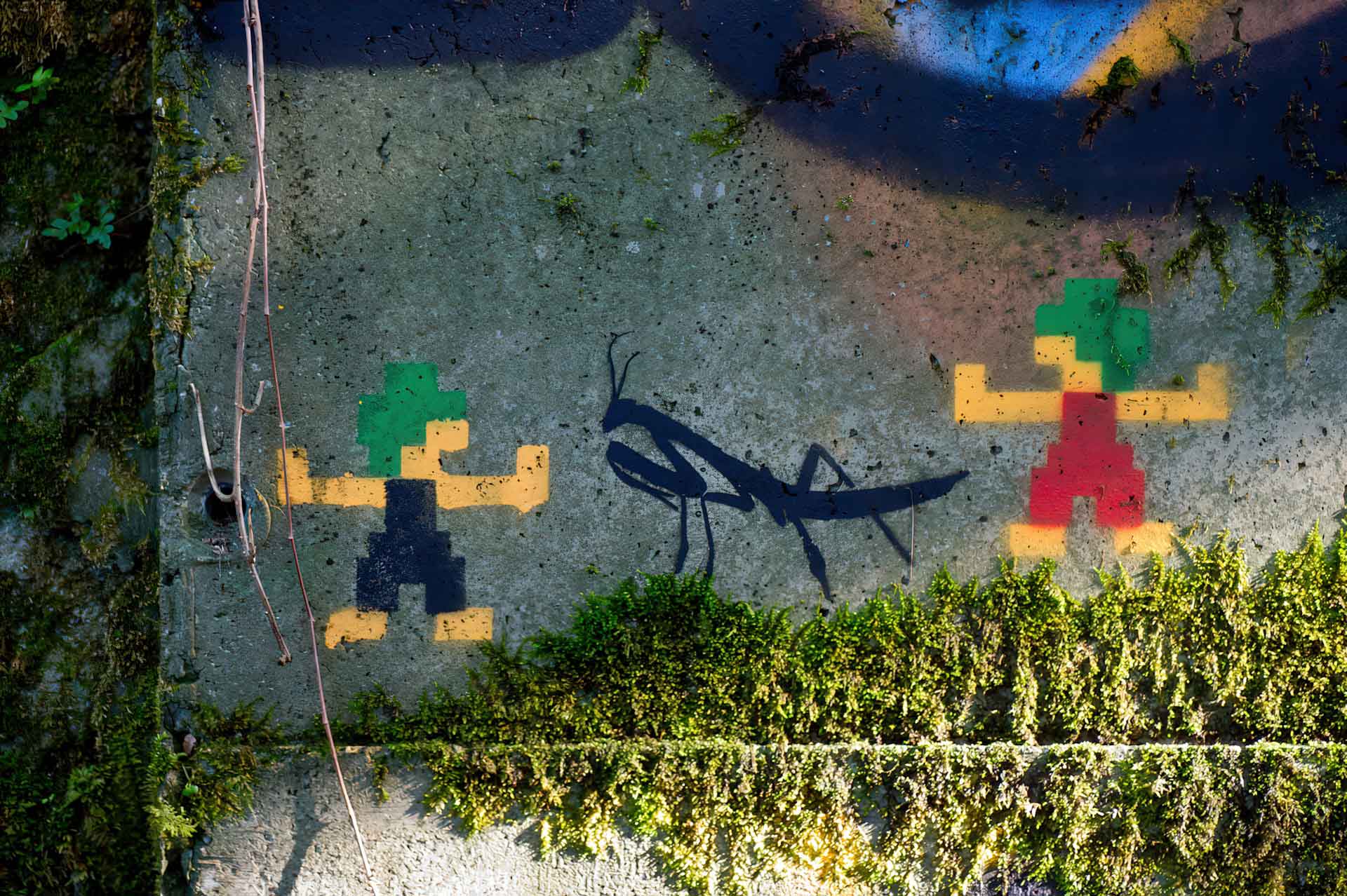 Pixel-style graffiti of Lemmings characters on a moss-covered concrete wall, with a black silhouette of a praying mantis between them.