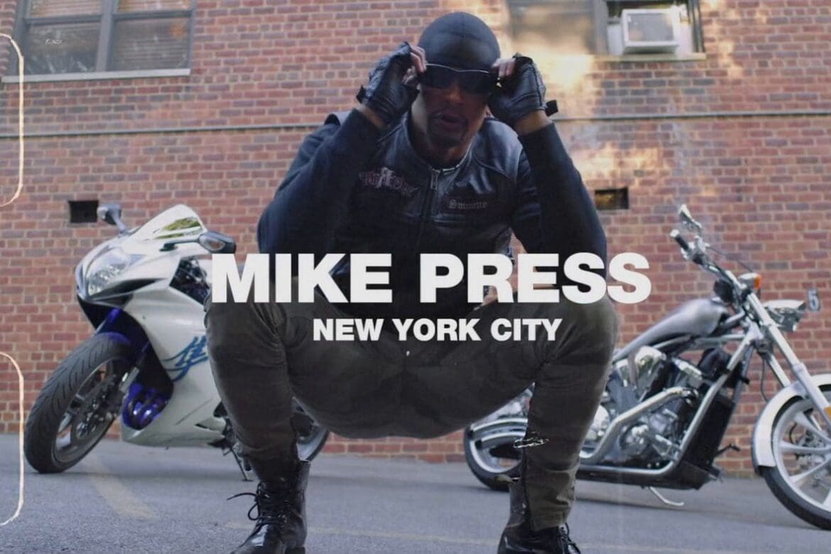 Mike Press crouching confidently in front of two motorcycles, wearing a leather jacket and sunglasses, set against an NYC brick wall.