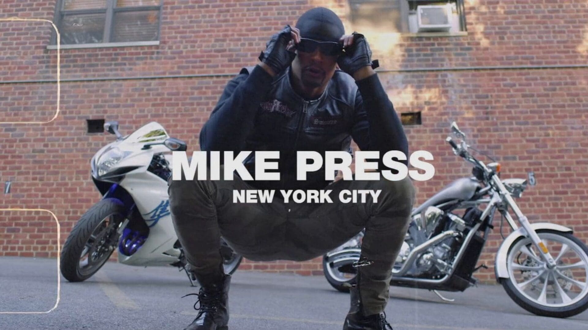 Mike Press crouching confidently in front of two motorcycles, wearing a leather jacket and sunglasses, set against an NYC brick wall.