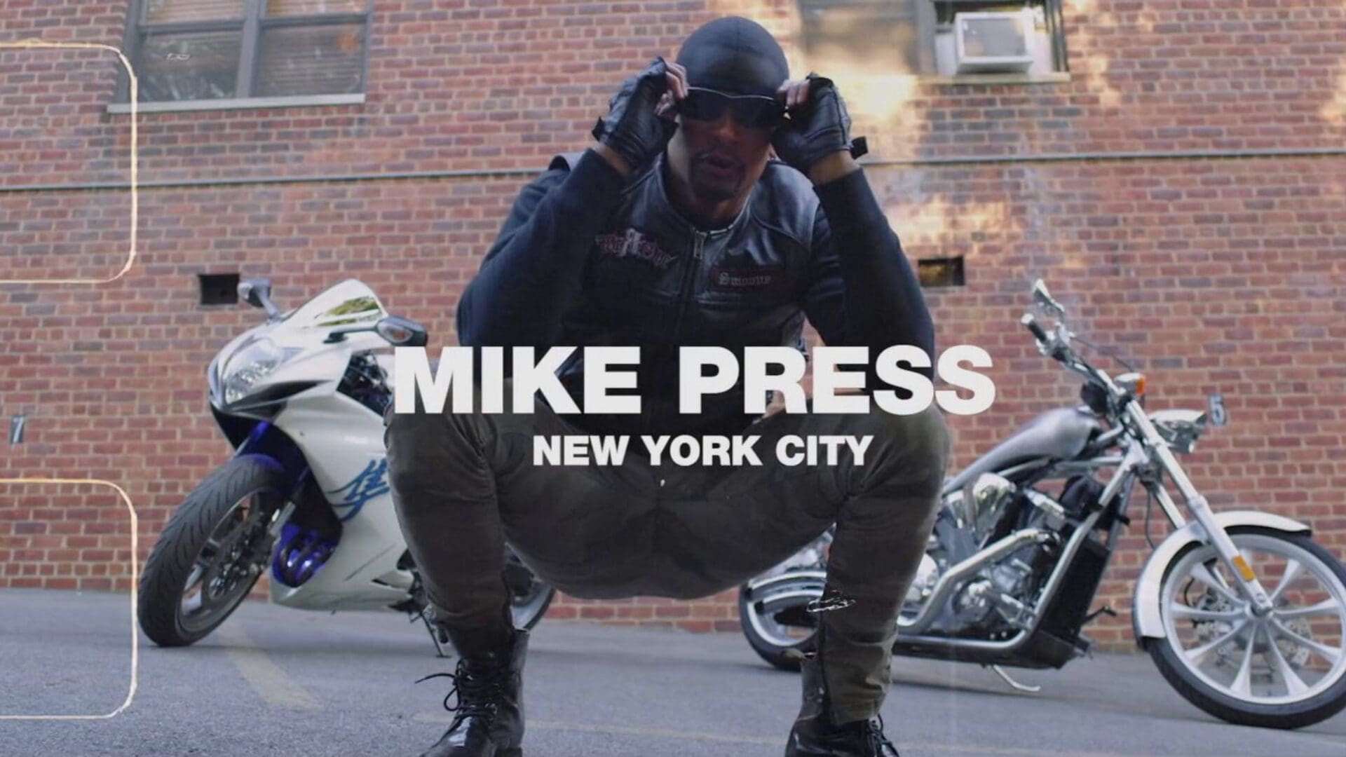Mike Press crouching confidently in front of two motorcycles, wearing a leather jacket and sunglasses, set against an NYC brick wall.