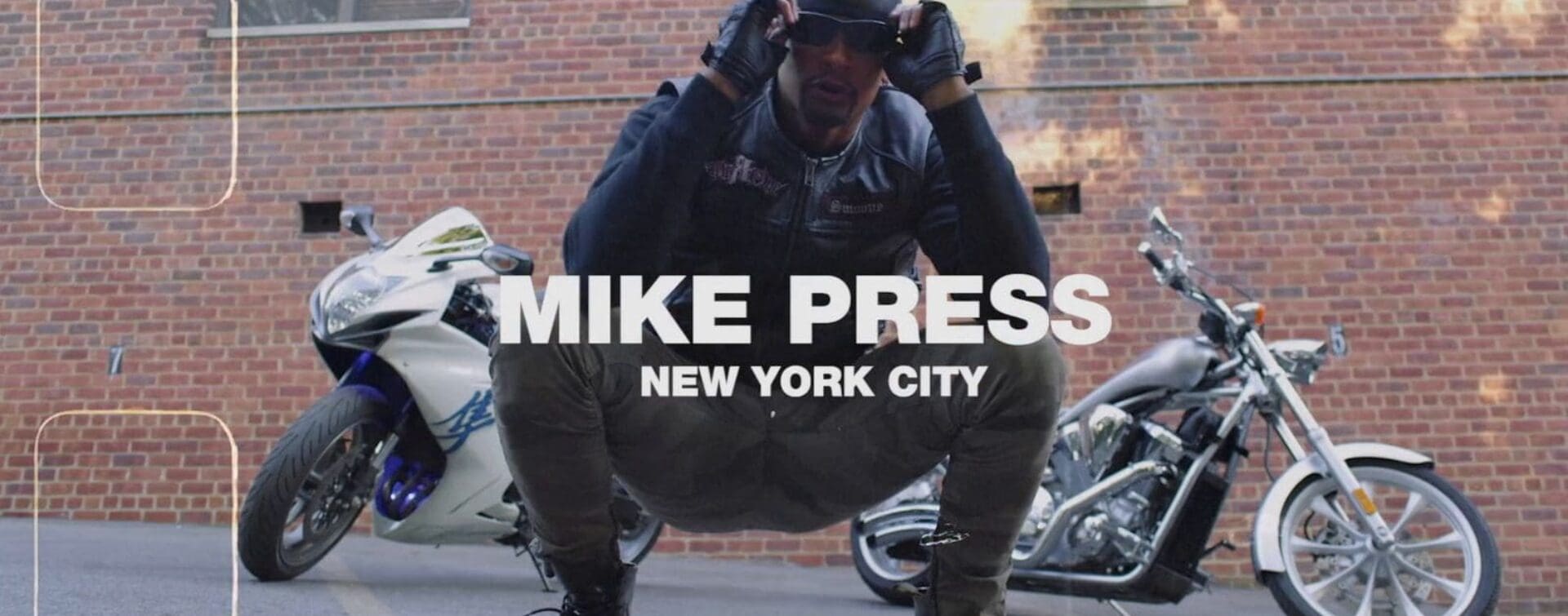 Mike Press crouching confidently in front of two motorcycles, wearing a leather jacket and sunglasses, set against an NYC brick wall.