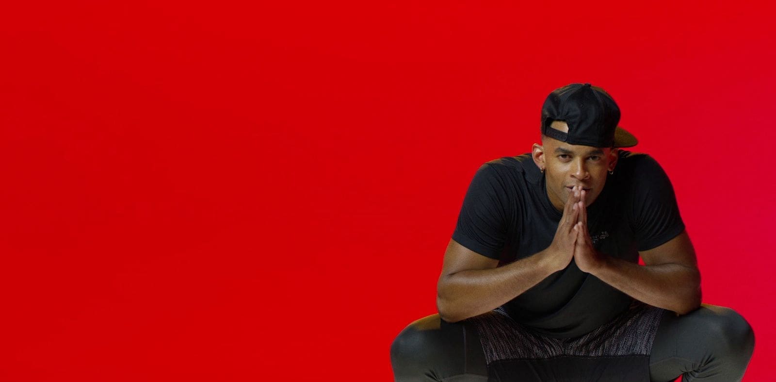 Mike Press crouching with hands clasped, wearing a backward cap, against a striking red background.