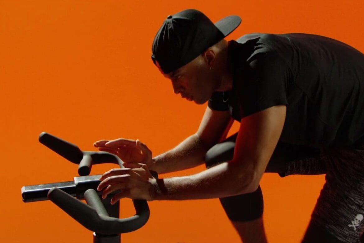 Mike Press riding a SoulCycle bike in a focused stance, set against a vibrant orange background.