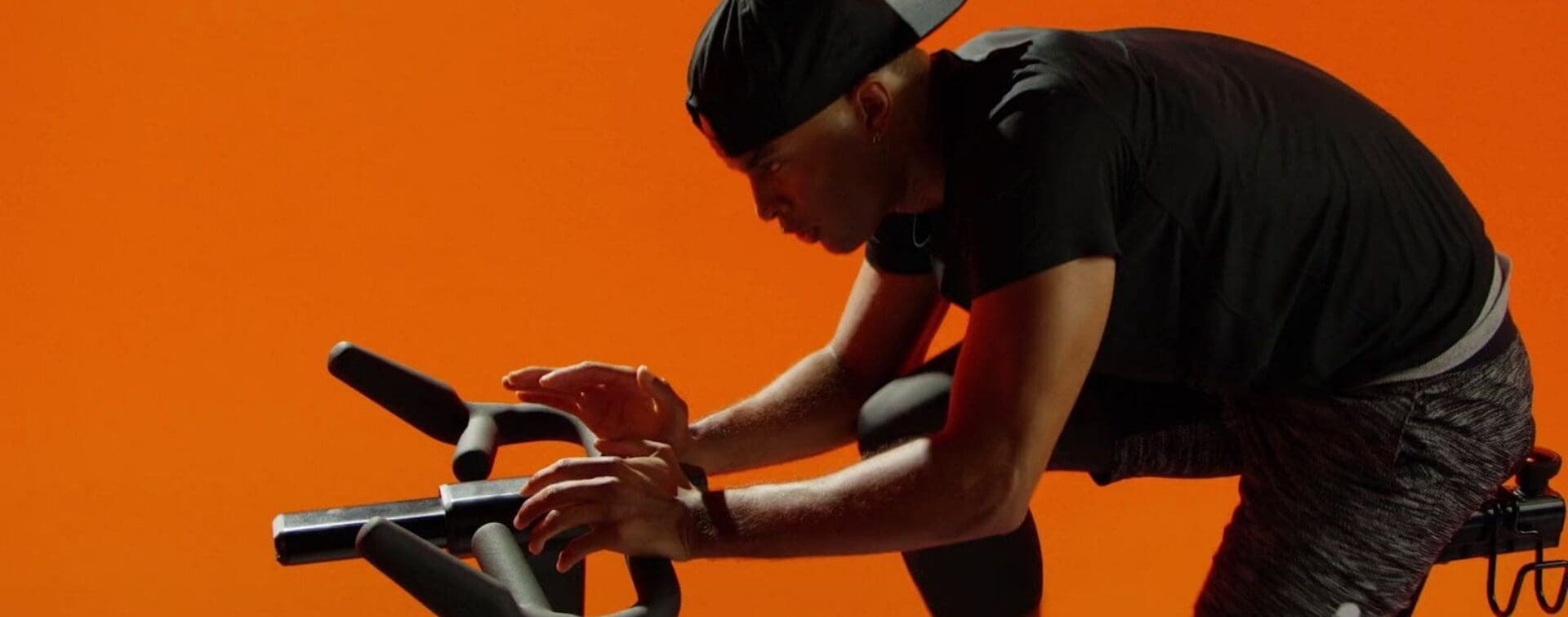 Mike Press riding a SoulCycle bike in a focused stance, set against a vibrant orange background.
