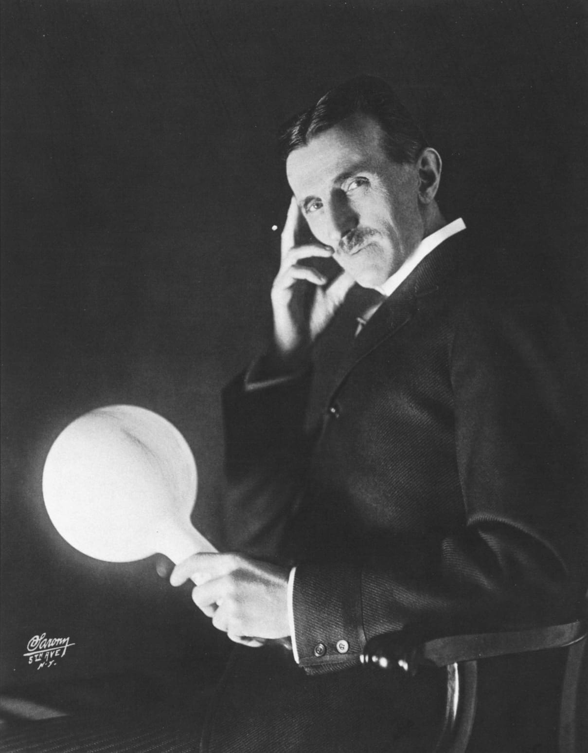 A portrait of Nikola Tesla holding a glowing, wireless light bulb, captured by photographer Napoleon Sarony around 1898.