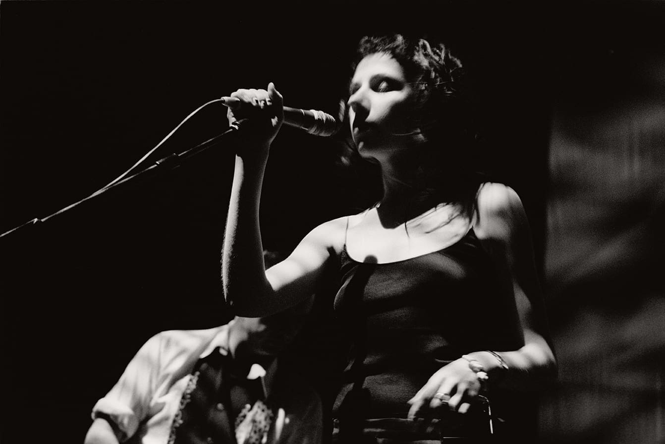 PJ Harvey performing live in 1998 with a microphone in hand, shot in black and white.