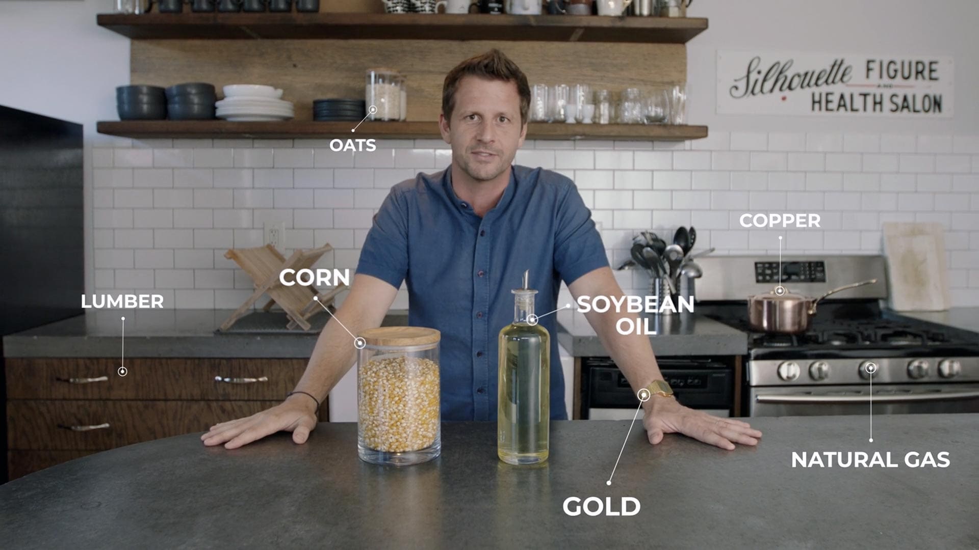 RIOT, NYC Creative Agency: Jamie McDonald standing in a kitchen with labeled commodities including corn, oats, soybean oil, lumber, gold, copper, and natural gas, illustrating futures trading concepts. Jamie McDonald standing in a kitchen with labeled commodities including corn, oats, soybean oil, lumber, gold, copper, and natural gas, illustrating futures trading concepts.