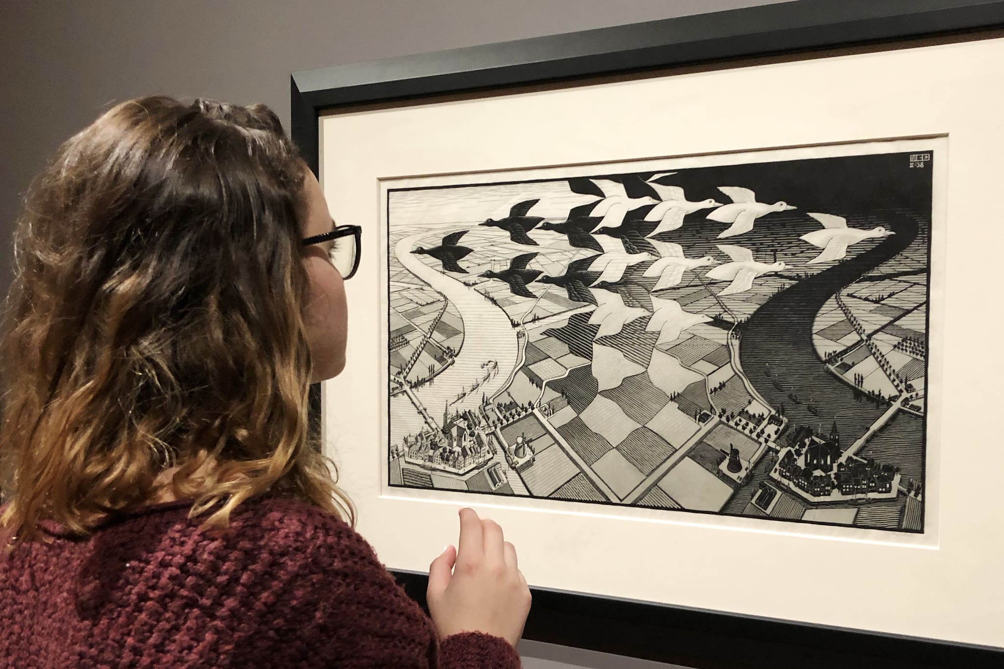 RIOT, NYC Creative Agency: A viewer observing M.C. Escher’s Day and Night artwork, depicting the transformation of birds over a divided landscape. A viewer observing M.C. Escher’s Day and Night artwork, depicting the transformation of birds over a divided landscape.