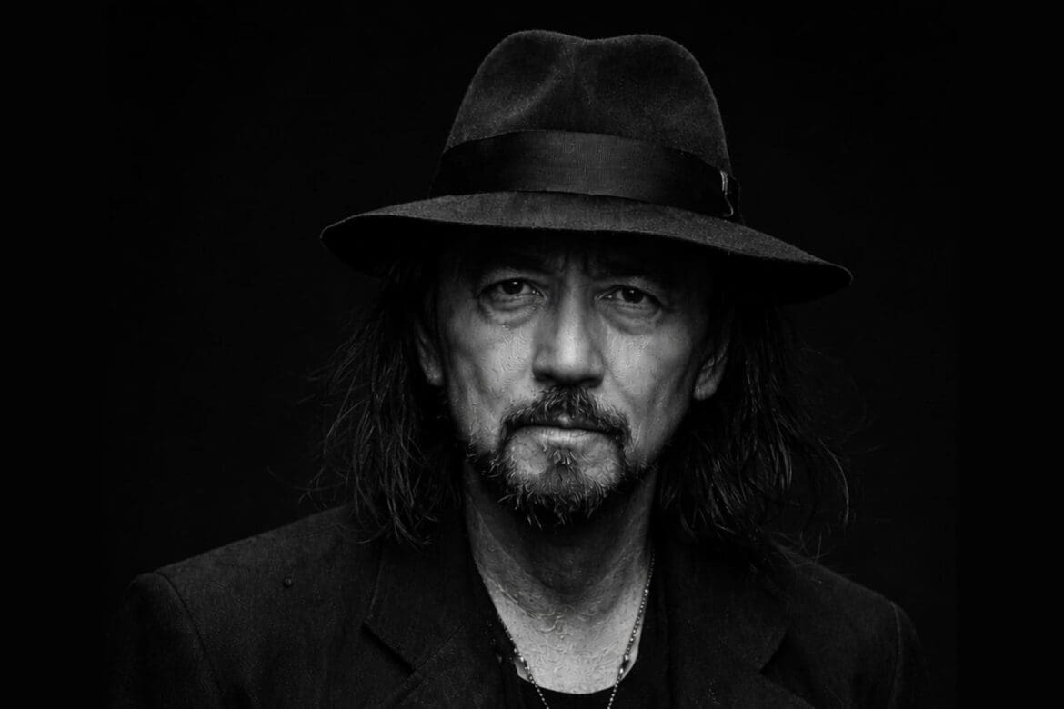 Black and white portrait of Yohji Yamamoto wearing a hat against a black studio background.
