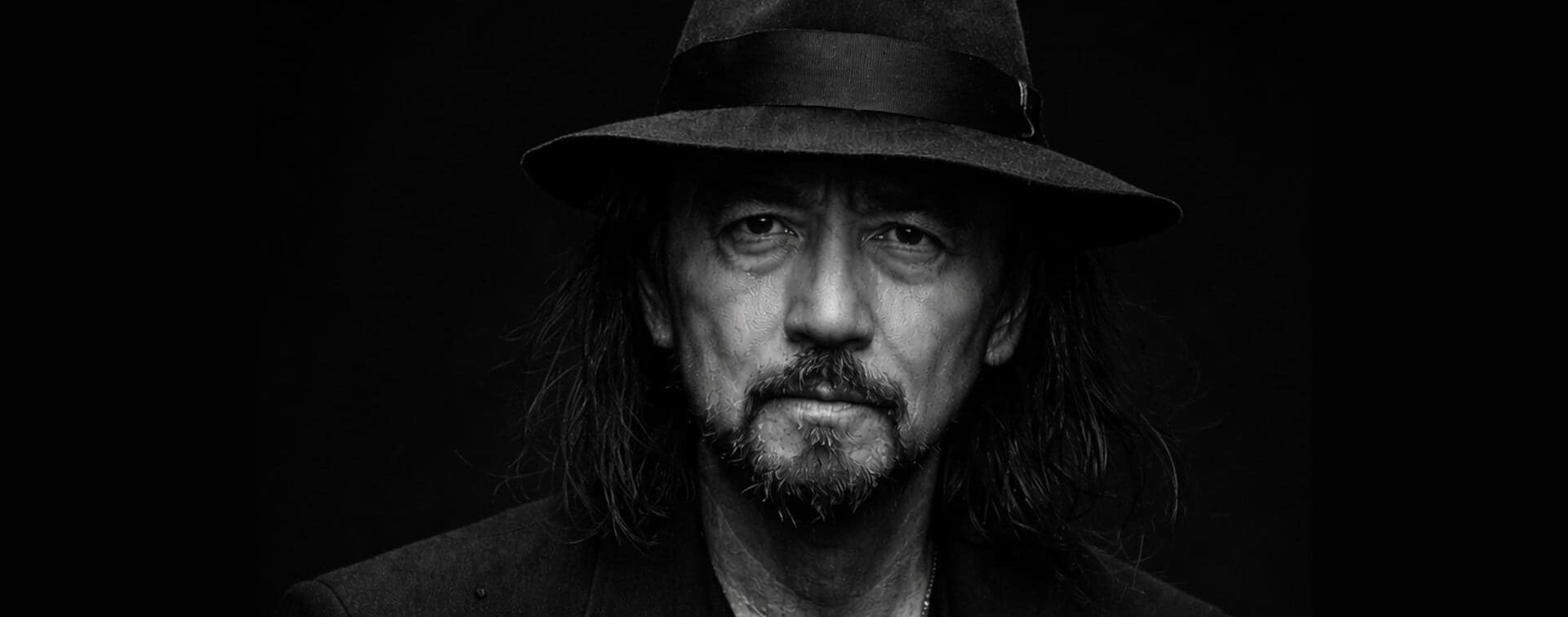 Black and white portrait of Yohji Yamamoto wearing a hat against a black studio background.